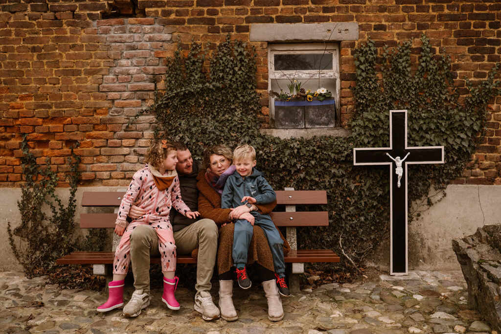 Gezin zit op bankje vlakbij hun huis. Typisch Limburgs aangezicht met een stenen muur, klimop en een kruis ernaast.