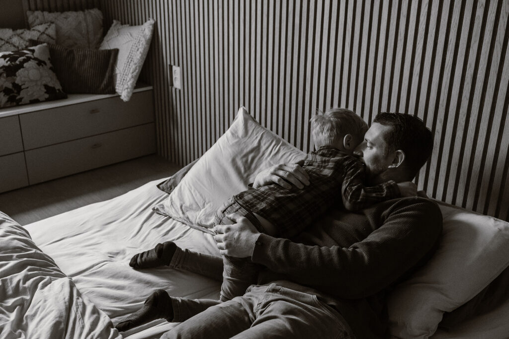 Een zwart/wit foto van een vader en zoon die elkaar een dikke knuffel op bed geven. 