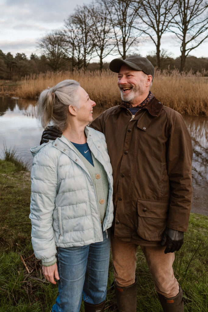 Een man en een vrouw staan bij de rivier de Regge in Ommen; de man heeft zijn arm om haar heen geslagen. Hij kijkt lachend weg en zij kijkt hem lachend aan. 
