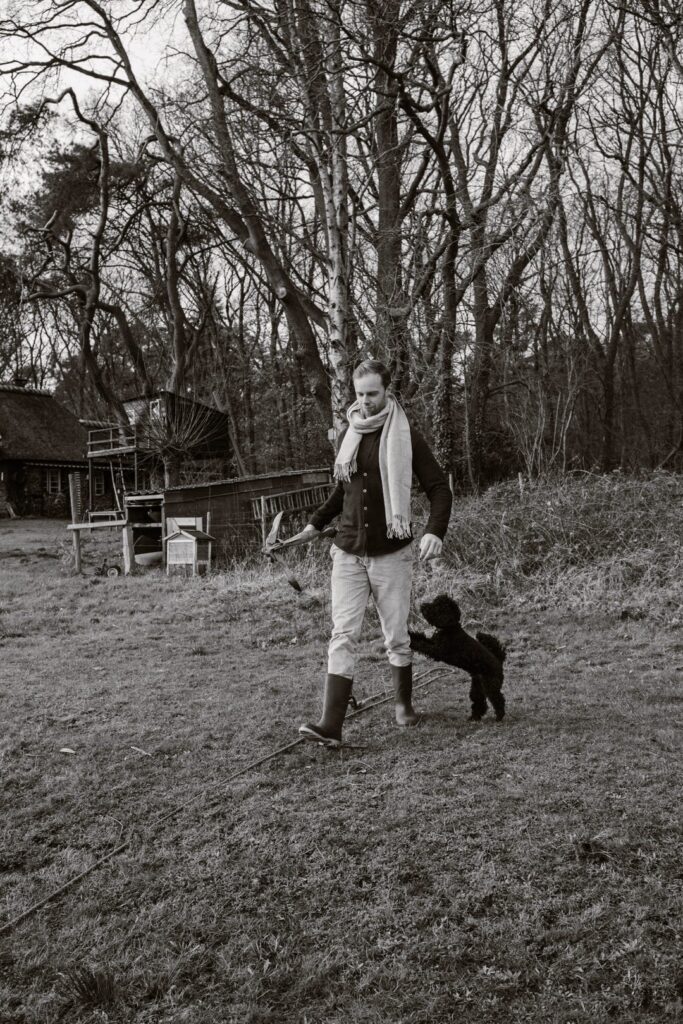 Een man in een modieuze outfit en leren laarzen loopt door een grasveld terwijl een vrolijke hond tegen hem opspringt